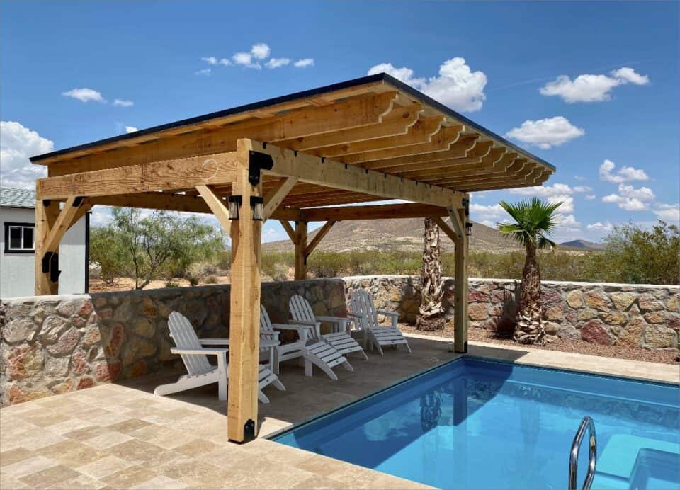 Custom wooden pergola with white Adirondack chairs next to a pool, surrounded by a desert landscape under a clear blue sky.