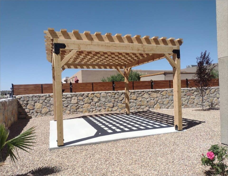 A wooden pergola from Wood You Too stands on a concrete base, casting shadows on the ground, with a high wooden fence and a stone wall in the backdrop under a clear blue sky.