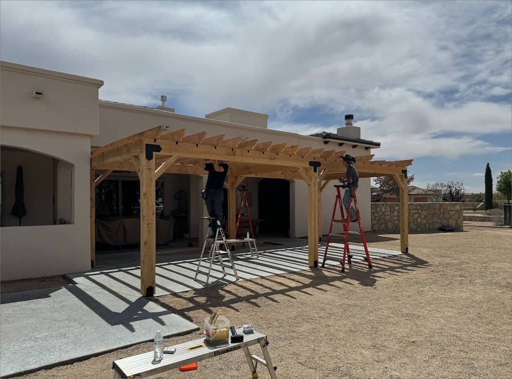 Two people constructing custom pergolas next to a residential building, with tools and ladders on a concrete patio under a cloudy sky.