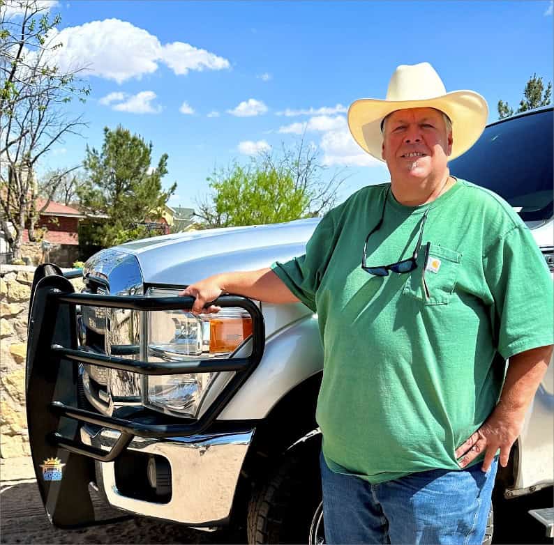 Man in a green shirt and cowboy hat standing beside a silver pickup truck with a Wood You Too custom pergola, under a clear blue sky.